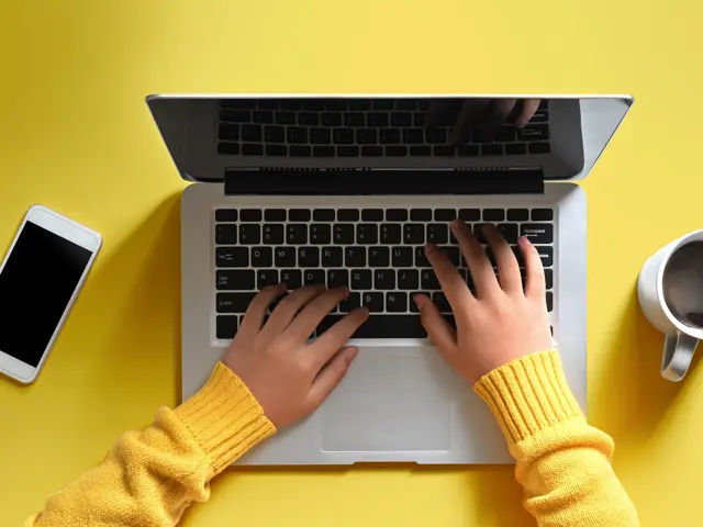 Birdseye view of someone using a laptop with a coffee and mobile phone next to them on a yellow table