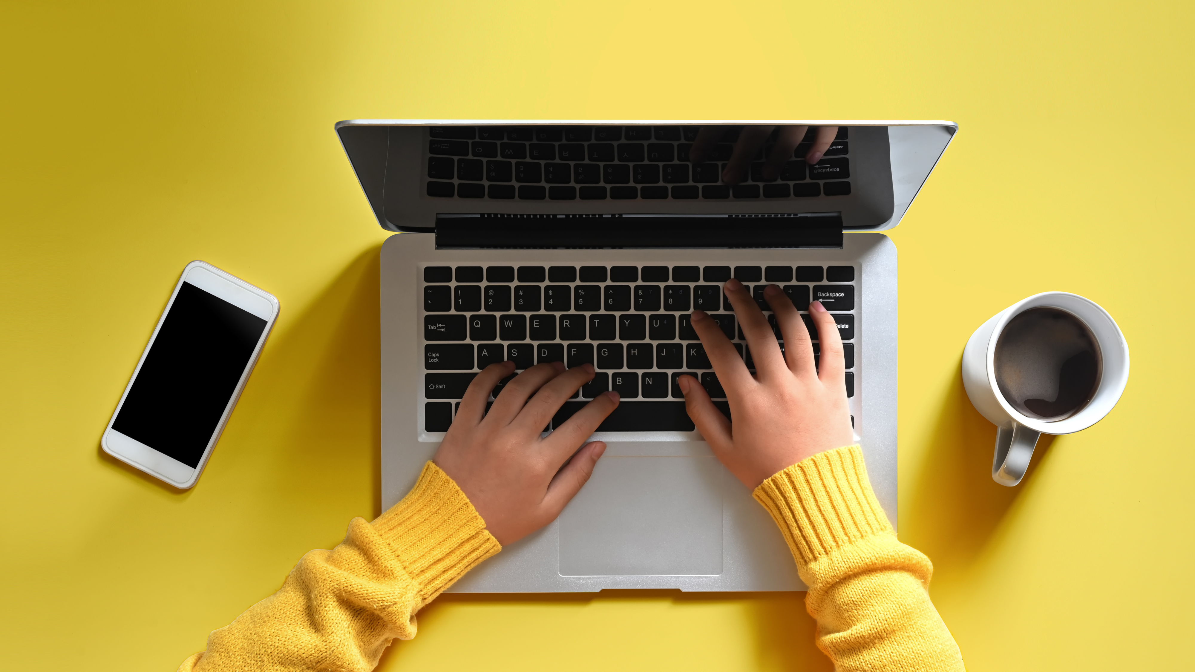 Birdseye view of someone using a laptop with a coffee and mobile phone next to them on a yellow table